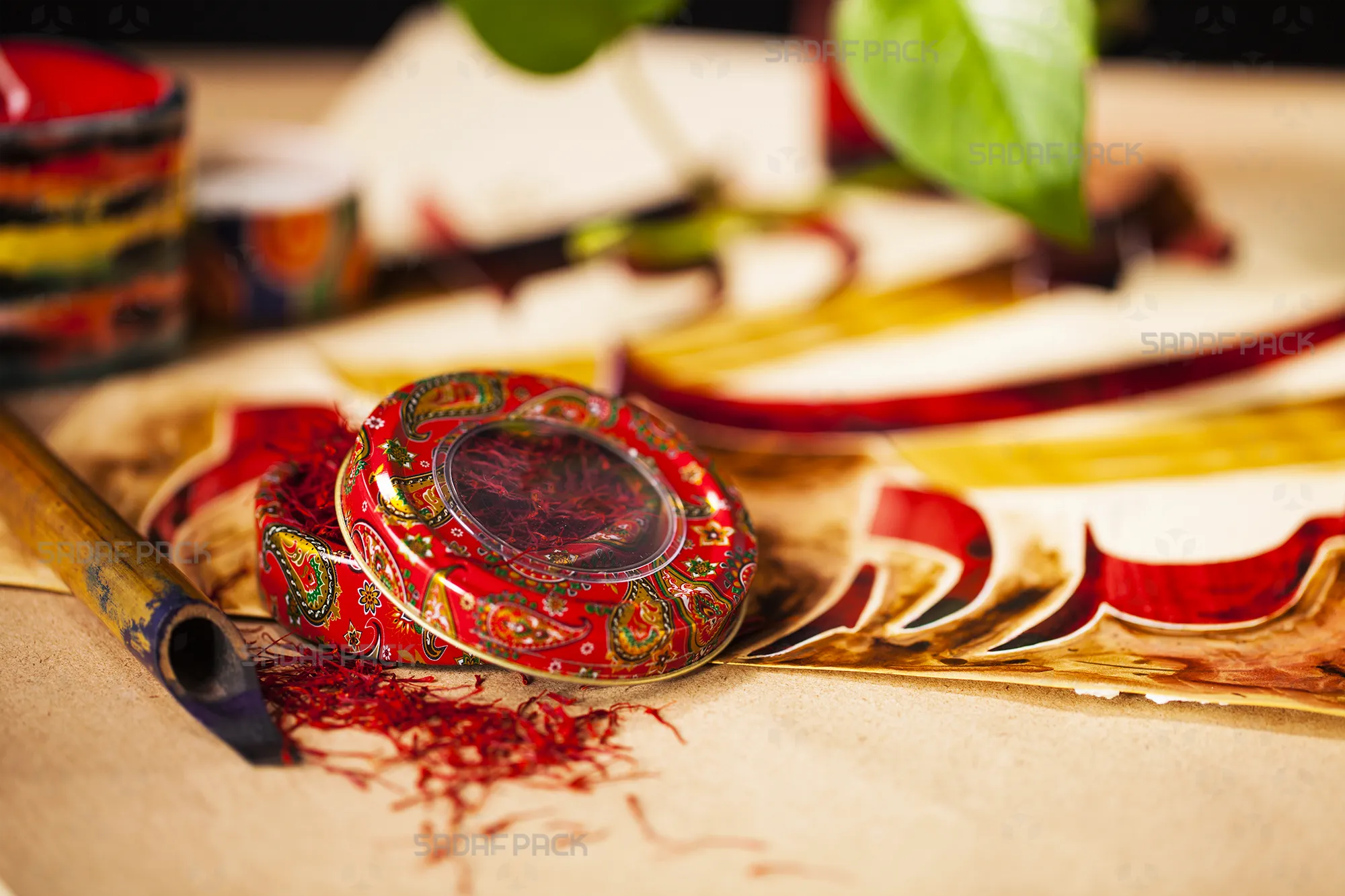 A Sadaf Pack 3 gram Metal Tin, featuring a vibrant Red Termeh (paisley) design, is displayed open with its clear-window lid set aside, revealing bright red saffron threads inside and scattered on a traditional parchment-like background.