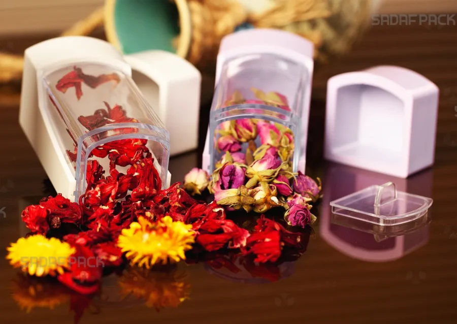 A pair of Sadaf Pack's elegant Negin poly-crystal containers, one with a white base and one with a lily base, showcasing vibrant dried hibiscus flowers and delicate rosebuds to highlight their suitability for luxury botanical packaging.