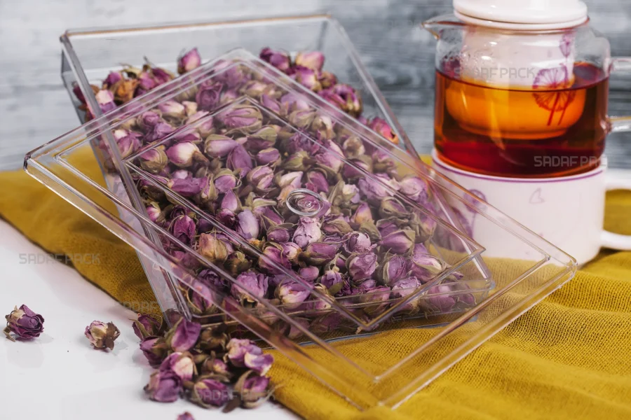 A Sadaf Pack 400 Crystal Rectangular container filled with dried rosebuds, shown from a three-quarter angle next to a glass teapot, highlighting its use for premium food products.