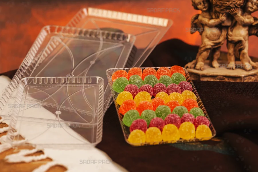 A clear Crystal Rectangle Tray from Sadaf Pack, elegantly filled with colorful, sugar-coated gumdrop sweets, showcasing its suitability for premium confectionery packaging. In the background, empty trays and lids are visible, highlighting the product's design.