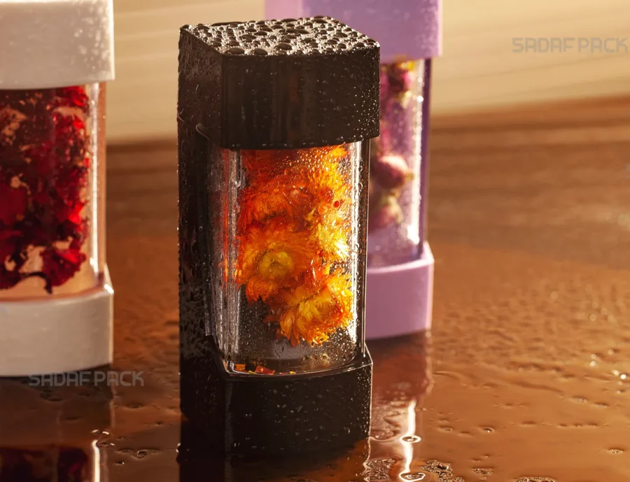 A striking close-up of the Sadaf Pack Negin container in black, its textured surface covered in fine water droplets. The container's transparent body showcases vibrant orange flowers inside, while the Negin White and Lily containers are softly blurred in the background, creating a luxurious and fresh aesthetic.