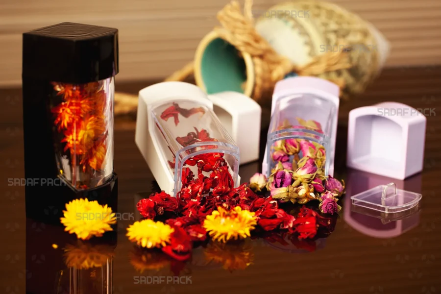 A stylish arrangement of Sadaf Pack Negin containers with black, white, and lily colored lids, displayed on a reflective surface and filled with vibrant dried rose buds, red hibiscus, and yellow flowers, showcasing their suitability for premium botanical products.