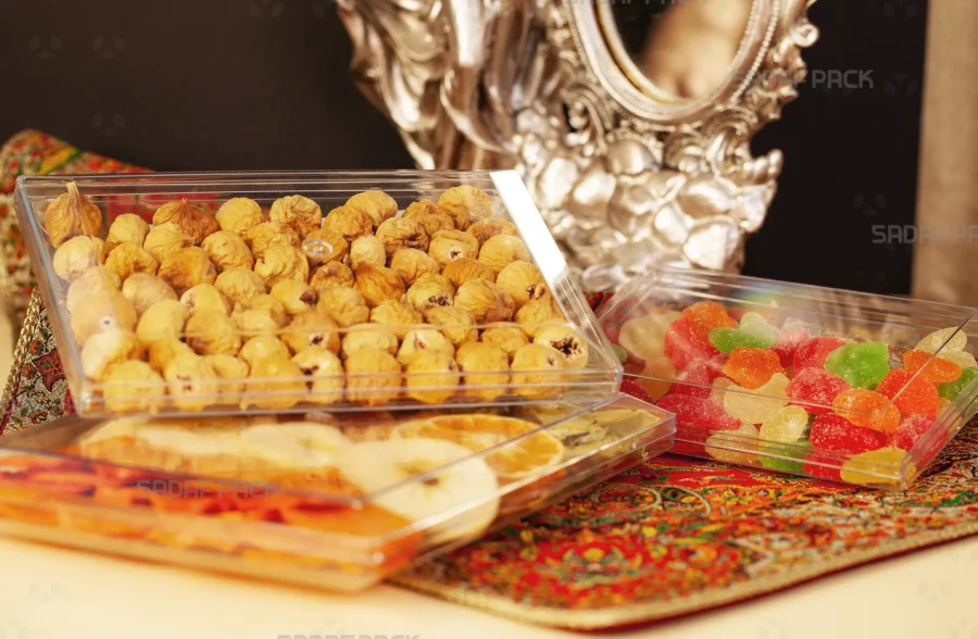 Three rectangular crystal containers by Sadaf Pack are displayed on an ornate tablecloth, filled with dried figs, dried orange slices, and colorful jelly sweets, showcasing their use for luxury food and confectionery packaging.