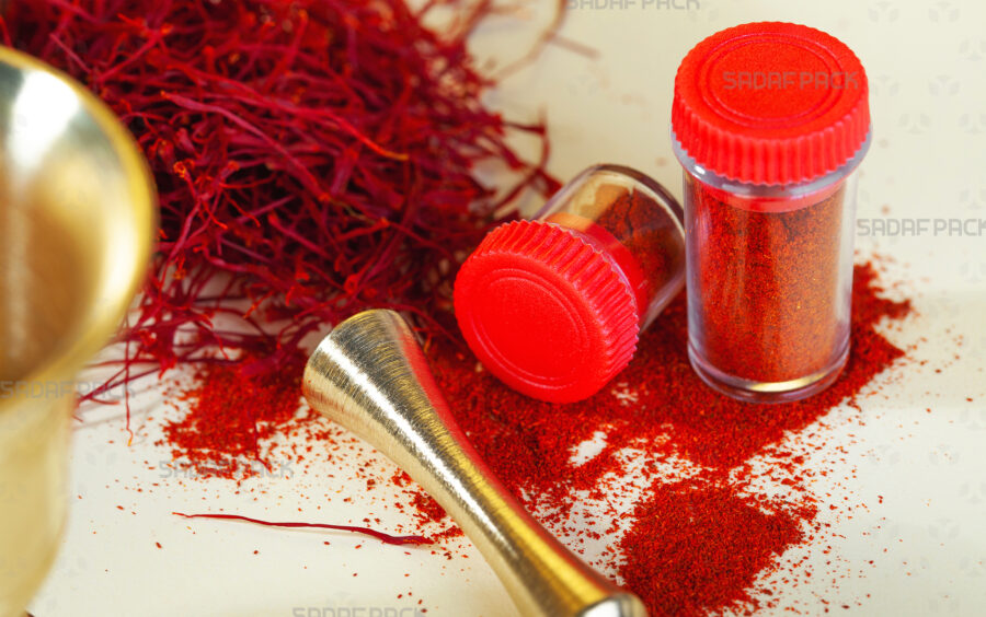 Two Sadaf Pack Powder Crystal Containers with red lids are displayed, one filled with rich ground saffron and the other empty, surrounded by vibrant red saffron threads and a traditional brass mortar and pestle to showcase their use for luxury spices.