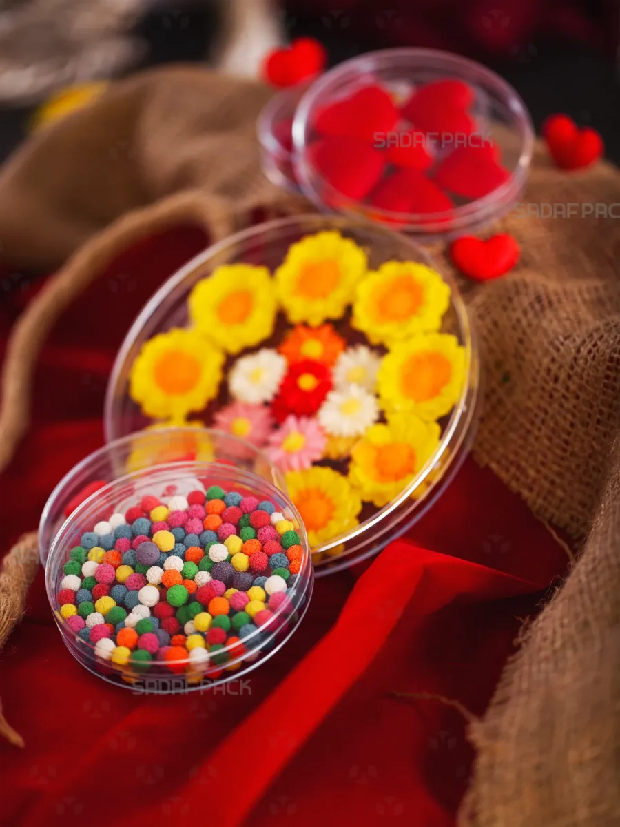 Three Sadaf Pack round deep crystal containers display a vibrant assortment of colorful sweets and candies. The largest container holds yellow and multi-colored flower-shaped gummies, while two smaller containers are filled with multi-colored sprinkle balls and red heart-shaped candies, showcasing their use for luxury gift packaging.