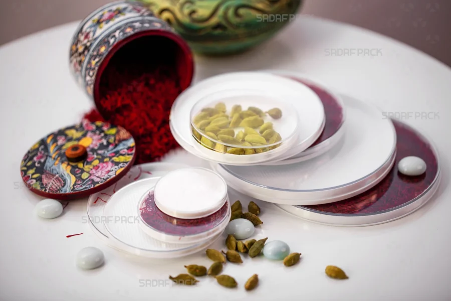 A collection of Sadaf Pack's elegant round containers with white bottoms and clear lids, beautifully displaying vibrant red saffron and green cardamom pods to highlight their premium packaging for luxury spices.