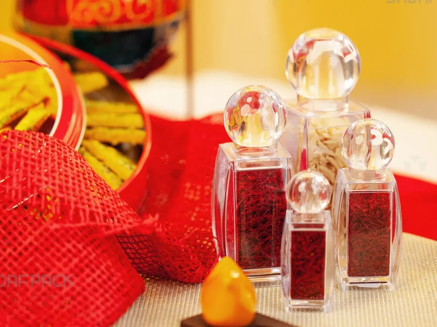 A luxurious collection of Sadaf Pack's sadaf crystal containers in various sizes, filled with vibrant red saffron threads and displayed in a warm, elegant setting suitable for premium food products.