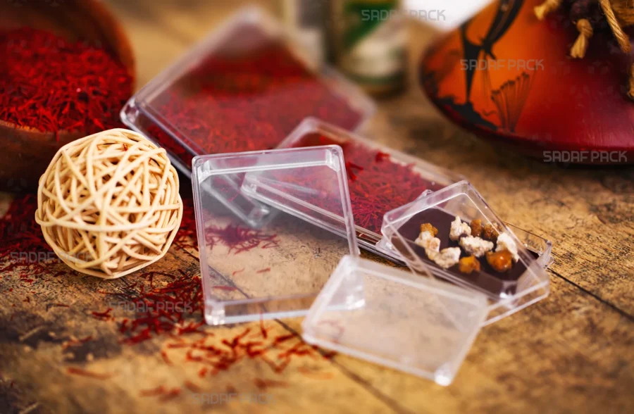 مجموعة من عبوات صدف باك الكريستالية المستطيلة الصغيرة والشفافة معروضة على طاولة خشبية. إحدى العبوات مملوءة بخيوط الزعفران الحمراء الزاهية وأخرى بالشوكولاتة الفاخرة، مما يوضح مدى تنوع استخداماتها في تغليف المواد الغذائية الفاخرة.