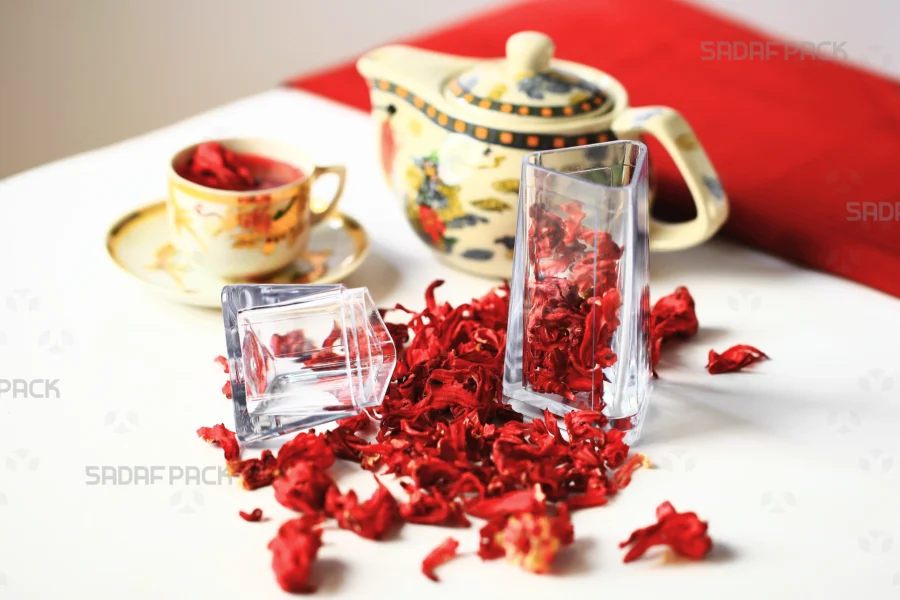 Sadaf Pack's Almas crystal container, filled and surrounded by vibrant red dried hibiscus flowers, displayed with a teapot and cup to showcase its suitability for premium herbal tea packaging.