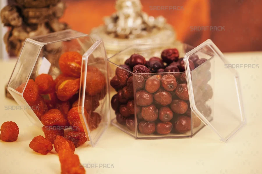 A Sadaf Pack short hexagonal clear polycrystal container filled with dried apricots and jujubes, elegantly arranged next to a decorative lit candle to showcase its suitability for luxury food packaging.